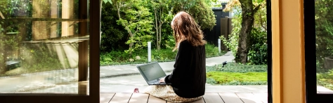 Woman wearing summer dress, sitting down outside on decking and typing on a laptop. Working from home business professional, employee, garden, green space, trees, wood doors, glass panes.