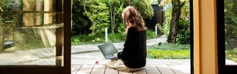 A woman sitting down on a garden patio wearing yellow summer dress. She is working and using her laptop outdoors in a lush green environment.