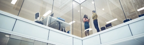 A pair of work colleagues talking with each other in a tall modern office building. Office has glass panes and working desks with bright lights and chairs.